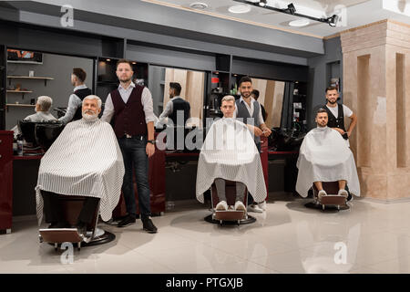 Three professional barbers standing near clients in barbershop. Male clients sitting in hairdresser chairs covered with striped haircut gowns. Men posing and looking at camera. Stock Photo
