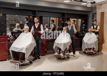 Male clients sitting in hairdresser chairs covered with striped haircut gowns. Three professional barbers standing near clients in barbershop. Men posing and looking at camera. Stock Photo