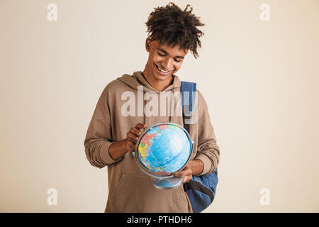 Portrait of a positive young afro american man dressed in hoodie carrying backpack isolated, holding earth globe Stock Photo