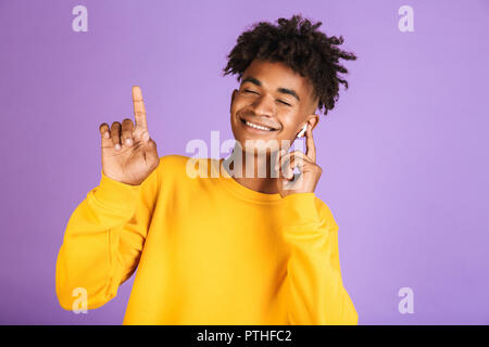 Portrait of happy african man having stylish afro hairdo dancing while listening to music via bluetooth earphone, isolated over violet background Stock Photo
