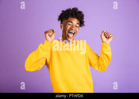 Portrait of happy african man having stylish afro hairdo dancing and singing, while listening to music via bluetooth earphone isolated over violet bac Stock Photo