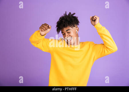 Portrait of young african american boy having stylish afro hairdo dancing and singing, while listening to music via bluetooth earbud isolated over vio Stock Photo