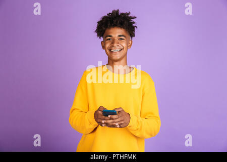 Portrait of teenager boy with afro hairdo smiling and holding smartphone, wearing bluetooth earphone isolated over violet background Stock Photo