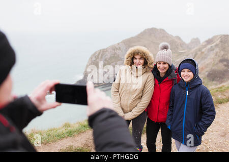 Man with camera phone photographing family in warm clothing on cliff overlooking ocean Stock Photo