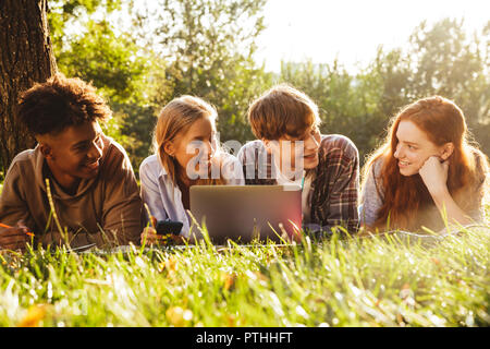 Group of excited multhiethnic students doing homework together at the park, using laptop computer, laying on a grass Stock Photo