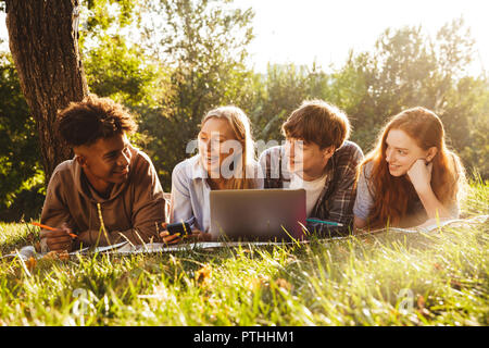 Group of joyful multhiethnic students doing homework together at the park, using laptop computer, laying on a grass Stock Photo