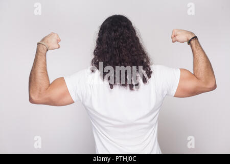 Back side of strong bodybuilder man in white t-shirt with long wavy hair, standing and posing with raised arms showing his biceps and muscular body. i Stock Photo