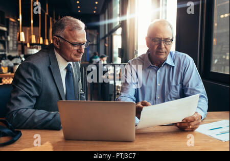 Senior businessmen discussing over a report while meeting in a cafe. Two mature businessmen working in cafe with laptop on table and looking at a docu Stock Photo