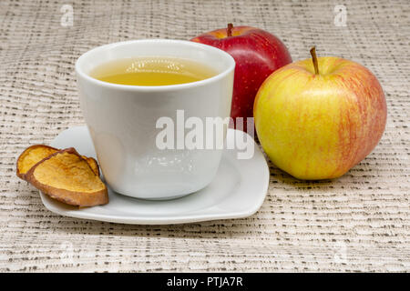 A cup of apple tea with two apples and dehydrated slices of the fruit with a patterned table cloth in the background Stock Photo