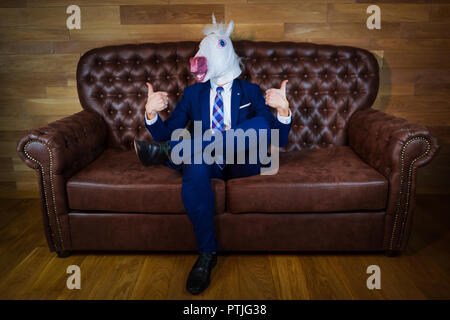 Funny unicorn in elegant suit sits on sofa like a boss and showing gesture thumbs up. Portrait of unusual man at home. Freaky guy in comical mask. Stock Photo