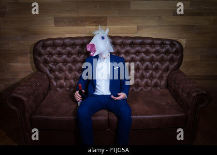 Funny unicorn in elegant suit sits on sofa with bottle of wine. Unusual man with headache at home. Freaky boss in comical mask. Stock Photo