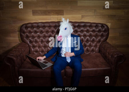 Strange man in elegant suit working at home office. Unusual young manager in comical mask on background of wooden wall. Funny unicorn sits on sofa. Stock Photo
