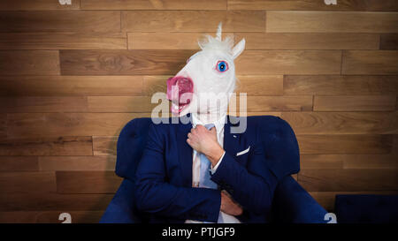 Portrait of unusual unicorn at home office. Freaky young manager in comical mask on background of wooden wall. Serious man in suit sits like boss Stock Photo