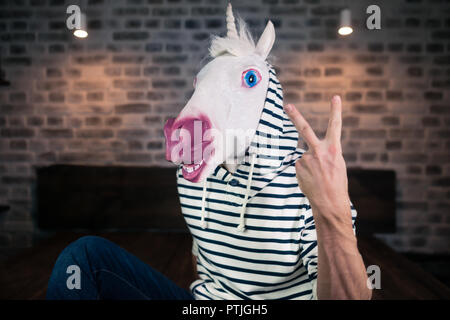 Freaky young man in comical mask sits on bed in stylish apartment. Funny unicorn in casual clothes is showing two fingers up gesture. Stock Photo