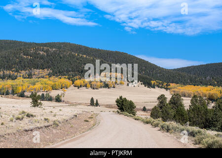 Autumn scene of a gravel road winding through grassy mountain meadows to hills and mountains covered in groves of golden aspen and green pines Stock Photo