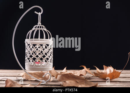 White decorative cage with candle hanging and burning on retro wooden desk with fallen dry leaves, black background. Autumn concept image Stock Photo