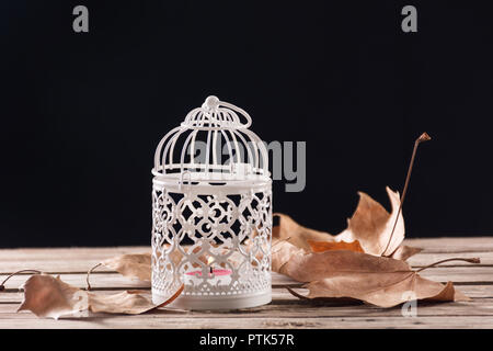 Decorative cage with candle burning on wooden retro desk with fallen dry autumn leaves. Black background. Close up, selective focus Stock Photo