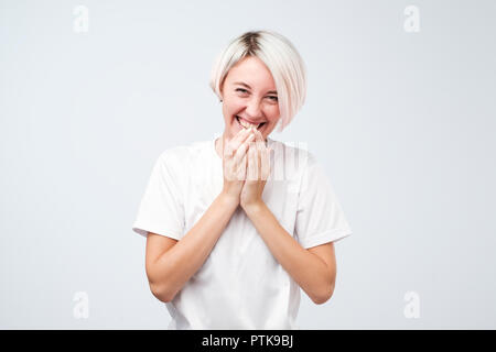 Good looking young female with dyed hair wearing white t-shirt giggles joyfully, covers mouth as tries stop laughing Stock Photo