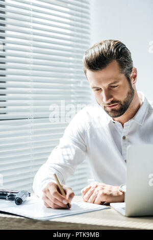handsome focused businessman doing paperwork at workplace with laptop and gun Stock Photo