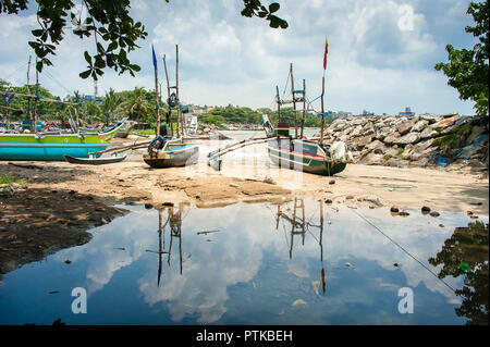 Traditional, painted, wooden fishing boats in the harbour. Colourful Oruwa catamarans sit on a sandy shore with cloudy sky reflections in sea water Stock Photo