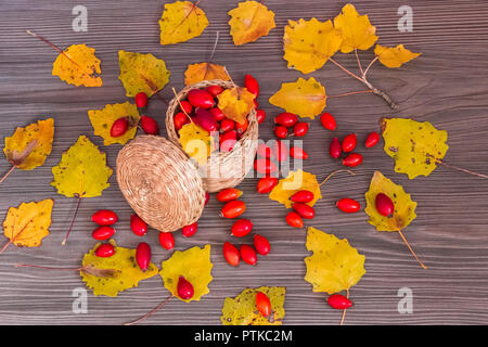 Rose hips in a basket and yellow leaves on a wooden surface, concept of autumn background Stock Photo