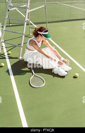 young woman in stylish white clothing and cap tying shoelaces on tennis court with racket and balls Stock Photo