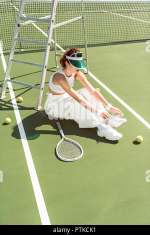 young woman in stylish white clothing and cap tying shoelaces on tennis court with racket and balls Stock Photo