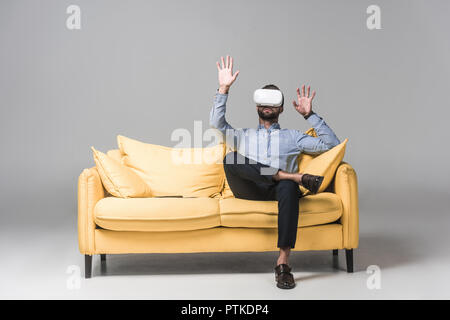 bearded man gesturing and using virtual reality headset and sitting on yellow sofa on grey Stock Photo
