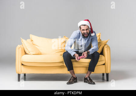 excited man in santa hat playing video game with joystick while sitting on sofa on grey Stock Photo