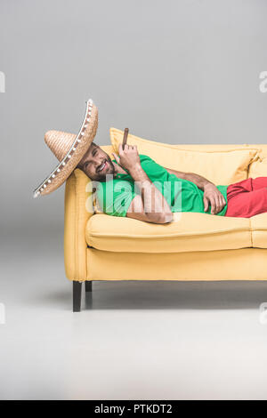 handsome cheerful man in mexican sombrero with cigar lying on yellow sofa on grey Stock Photo