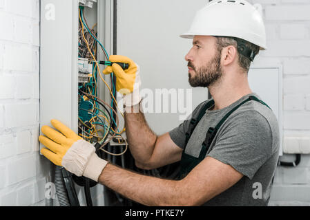 side view of handsome bearded electrician repairing electrical box and using screwdriver in corridor Stock Photo