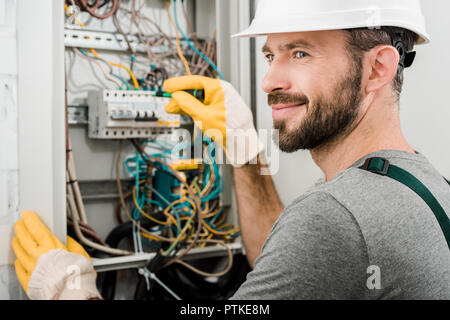handsome cheerful electrician repairing electrical box and using screwdriver in corridor Stock Photo