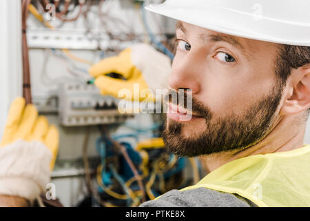 handsome bearded electrician repairing electrical box, using screwdriver in corridor and looking at camera Stock Photo