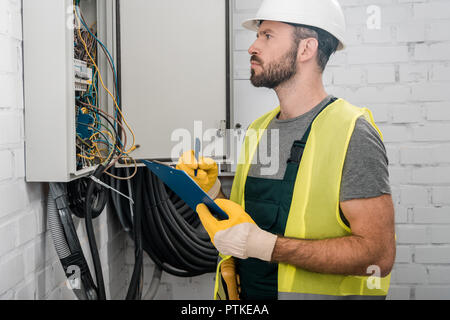 side view of serious electrician holding clipboard and checking electrical box in corridor Stock Photo