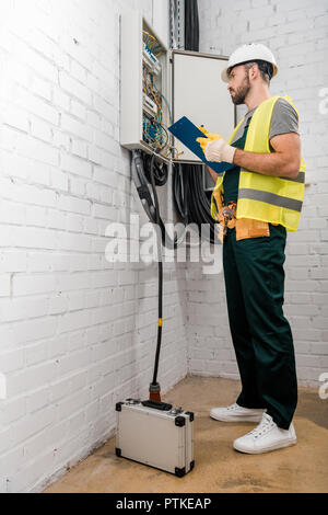 low angle view of handsome electrician holding clipboard and checking electrical box in corridor Stock Photo