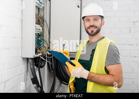 smiling handsome electrician holding clipboard near electrical box in corridor and looking at camera Stock Photo