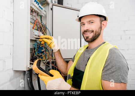 smiling handsome electrician checking electrical box with multimetr in corridor and looking at camera Stock Photo