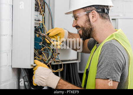 side view of smiling handsome electrician repairing electrical box with pliers in corridor Stock Photo