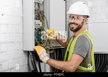 smiling handsome electrician repairing electrical box with pliers in corridor and looking at camera Stock Photo