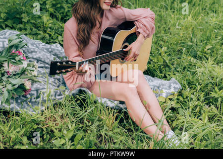 partial view of woman playing acoustic guitar while resting on blanket with bouquet of flowers in park Stock Photo