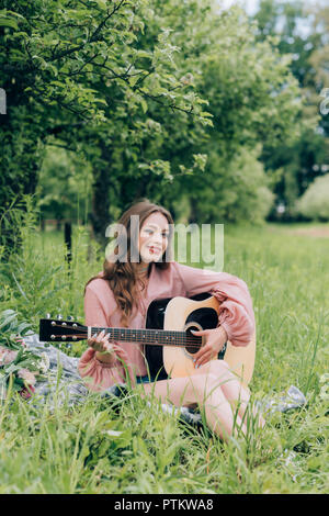 young smiling woman with acoustic guitar resting on blanket with bouquet of flowers in park Stock Photo