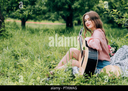 side view of young woman with acoustic guitar resting on blanket with bouquet of flowers in park Stock Photo