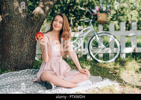 smiling pensive woman with ripe apple in hand resting on blanket under tree at countryside Stock Photo