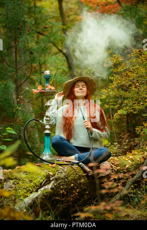 Young redhead woman in hat smoking Hookah in forest Stock Photo