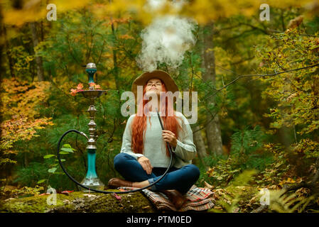 Young redhead woman in hat smoking Hookah in forest Stock Photo