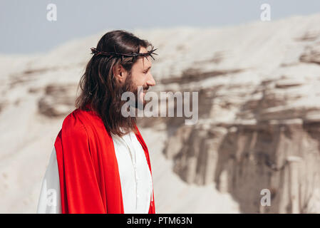 side view of smiling Jesus in robe, red sash and crown of thorns looking away in desert Stock Photo