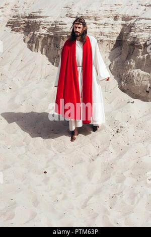 Jesus in robe, red sash and crown of thorns walking in desert Stock Photo