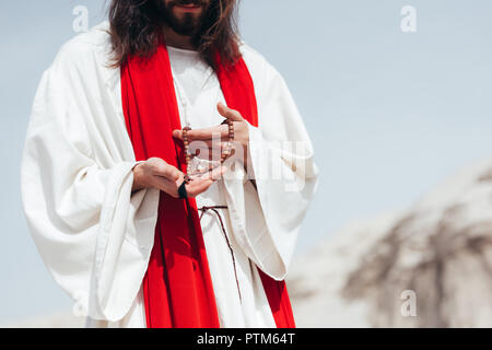 cropped image Jesus with long hair in robe and red sash holding wooden rosary in desert Stock Photo