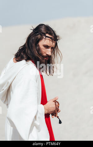 side view of Jesus in robe and red sash holding wooden rosary in desert Stock Photo