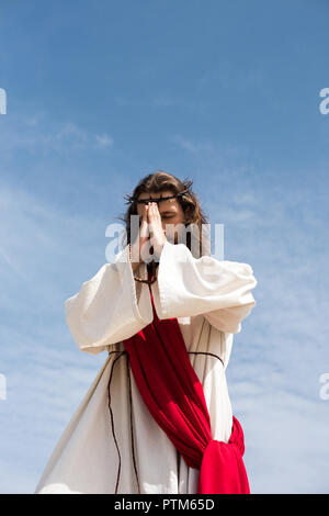 low angle view of Jesus Christ in robe, red sash and crown of thorns holding rosary and praying against blue sky Stock Photo